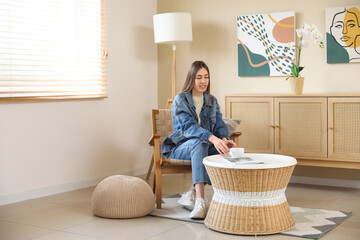 Canvas Print - Young woman having cup of tea while sitting in armchair near coffee table in room