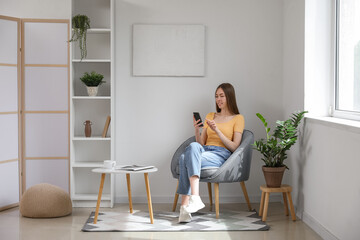 Wall Mural - Young woman using mobile phone while sitting in armchair in living room