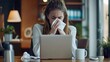© Oulaphone - Sick young woman works in the office at a desk, sits in a chair in front of a laptop, sneeze or wipes his nose with a tissue.