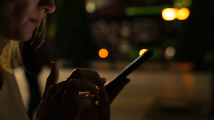 Close-up of a woman with a mobile phone in her hands on the street in the dark, posting messages on social networks, shopping online, sending text messages.