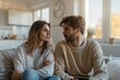 © Milos - A couple sits on a couch at home, both deep in thought, staring towards each other with expressions that indicate a meaningful and possibly emotional conversation.