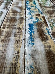 A detailed close-up shot of a weathered rustic wooden table surface, showing layers of worn paint in blue, white, and natural wood tones. The texture and distressed look of the wood highlight its age