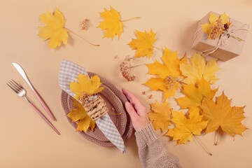 Wall Mural - Autumn table setting on beige background. Womens hand holding oval plates, cutlery, checkered napkin, gift box and dry yellow leaves. Autumn holiday mood, Halloween, Thanksgiving concept. Top view