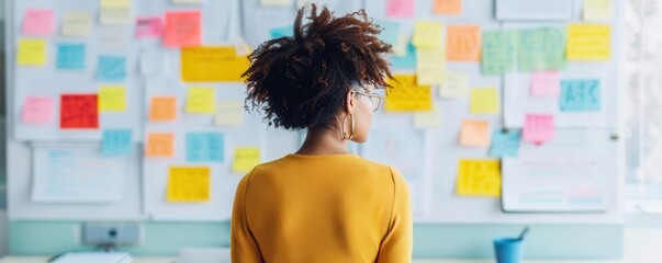 A thoughtful woman observes a colorful wall of sticky notes, reflecting on ideas and plans in a creative workspace.