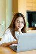 © Ekkasit A Siam - Asian woman in glasses and white shirt working on a laptop at a wooden table, hand on chin. Background includes a coffee cup. Captures thoughtful, professional moment of remote work
