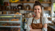 © Alloca Design Studio - A smiling young Caucasian woman with long brown hair in a bun, wearing a brown apron, standing in a bakery with a bright display case filled with donuts.