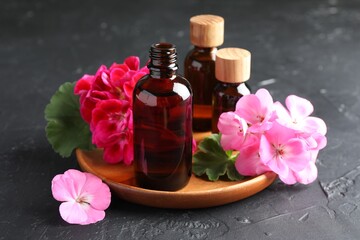 Bottles of geranium essential oil and beautiful flowers on black table, closeup