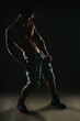 © qunica.com - Young shirtless man in denim shorts and boots posing confidently in a dark setting with dramatic lighting highlighting his muscles.