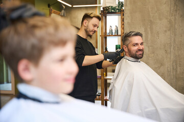 Caring father smiling watching qualified barber cutting his son hair