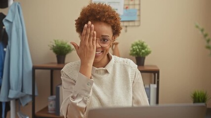 Sticker - Cheerful african american woman confidently covers one eye with hand, wearing glasses, sitting at home table and flashing a fantastic smile