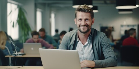 Wall Mural - A young man in an open office, smiling at the camera, sitting at a desk with a laptop computer. Workspace featuring contemporary design elements.