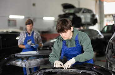 Young professional auto body technician in blue overalls and casual green hoodie, engaged in restoring damaged cars, expertly sanding removed bumper with sandpaper to smooth surface before painting