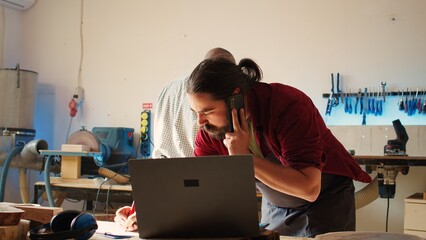 Sticker - Cabinetmaker receiving telephone call from client commissioning furniture, writing down info. Carpenter in assembly shop drawing schematics, talking on smartphone with customer about order, camera B