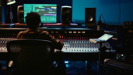 Wall Mural - African american sound designer editing audio recordings in control room, working with switchers and faders on mixer console. Music producer creating new tunes and tracks. Camera B.