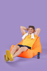 Canvas Print - Handsome young man with sports bottle sitting in beanbag chair on lilac background