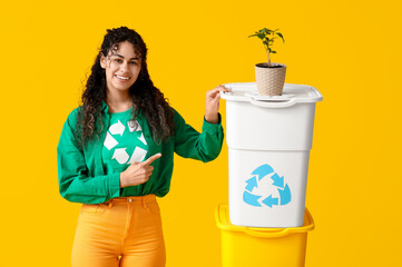 Beautiful young African-American woman in t-shirt with recycling symbol pointing at houseplant and trash bins on yellow background