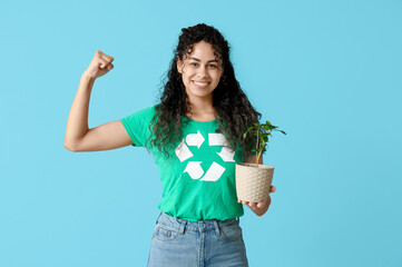 Beautiful young African-American woman in t-shirt with recycling symbol holding houseplant and showing muscles on blue background