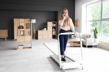 Wall Mural - Young businesswoman talking by mobile phone on treadmill in office