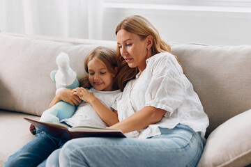 Wall Mural - Mother and daughter bonding on couch reading a book together while holding a teddy bear