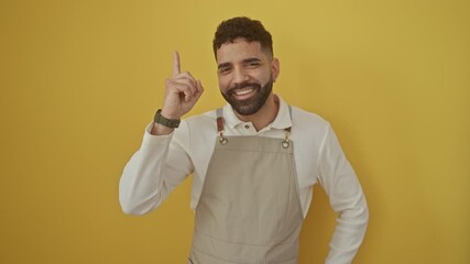 Sticker - Young, happy hispanic man wearing apron, standing over isolated yellow background, pointing up with one finger as a gestural sign of success, excitement, and a brilliant idea number one