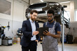 © sofiko14 - Mechanic in gray overalls showing tablet to businessman in suit inside car repair shop. Discussing vehicle maintenance details. Car on lift in background. Professional cooperation, automotive industry