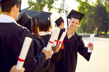 Happy college, university or high school student boy at his graduation. Young man in black cap and gown with diploma in hand looks back and smiles at camera while standing in line with other students