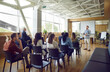 © Studio Romantic - Young berded man speaking on a business training standing in office during a conference or seminar for a group of diverse people company employees sitting on chairs in meeting room.