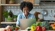 © olga_demina - African American woman working on her laptop surrounded by fresh vegetables in a modern kitchen. Nutrition and healthy eating concept.