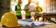 © inthasone - Close-up of a yellow helmet on a construction site table with a blurred engineer and architects in the background, holding paper and a laptop computer working together on a new project plan.