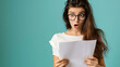 © Gita - A young woman wearing glasses with a surprised expression holding a document against a light blue background, capturing a moment of astonishment and disbelief.