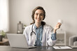 © fizkes - Portrait of young intern, internist woman in white coat smile, look at camera seated at desk. Workflow of female GP using laptop at workplace, provide professional consultation to patients remotely