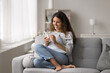 © fizkes - Smiling Latina woman resting on cozy sofa with smartphone, making call, check personal organizer using mobile application, enjoy remote communication with friend, ordering delivery services at home