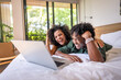 © lordn - A young couple of African descent is lying on a bed in a modern, sunlit bedroom, smiling and looking at a laptop screen. They appear to be enjoying a pleasant moment together, relaxed and happy.