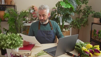 Wall Mural - Bearded man with grey hair working on a laptop in a colorful flower shop surrounded by plants and bouquets.