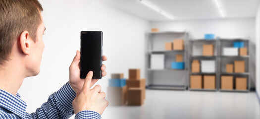 Man with phone is standing in warehouse. Guy inside storage room. Businessman in storage room with boxes. Man uses application for accounting in warehouse. Cropped contractor conducts storage audit