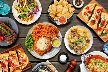 Poster - Table scene of various delicious foods. Top down view on a dark wood background. Pastas, flatbreads, tacos, steak and restaurant theme dishes.