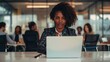 © ImronDesign - Attractive african young confident businesswoman sitting at the office table with group of colleagues in the background, working on laptop computer - generative ai