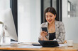 © PRIME STOCK LAB - Asian businesswoman using smartphone at office workstation, with a desktop computer and financial reports on her desk.