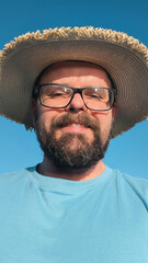 Poster - Selfie portrait of handsome bearded man with glasses, straw hat and blue shirt making facial expressions, isolated on blue background