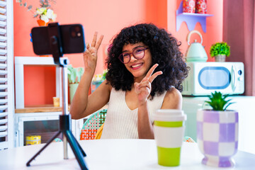 Wall Mural - Beautiful young hispanic woman with curly hair portrait at home - Happy teenager influencer streaming live on social media with her mobile phone