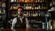 © Luiri Art - Young male bartender standing behind the bar counter in a dimly lit pub with shelves of liquor bottles in the background, wearing a brown vest and white shirt, looking confidently at the camera