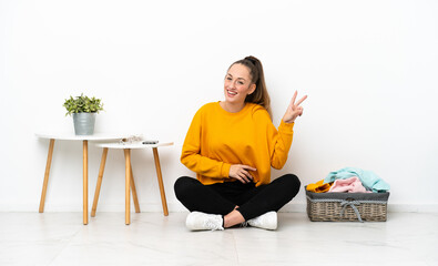 Wall Mural - Young caucasian woman folding clothes sitting on the floor isolated on white background smiling and showing victory sign