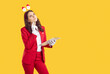 © Studio Romantic - Event manager writing checklist for Christmas party. Beautiful woman in red suit and Santa headband standing on yellow copy space studio background, holding clip board and thinking with hand on chin