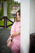 © keleny - A young woman with long brown hair is wearing a pink shirt and is leaning against a white post