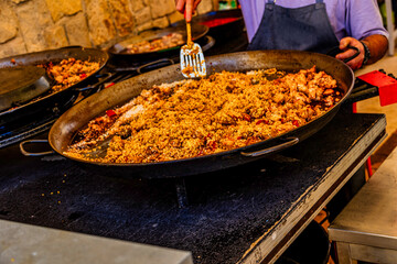 A  metal pan filled with a seasoned dish, possibly paella, is being cooked on a stovetop with a person stirring it.