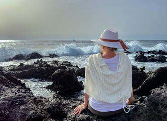 Canvas Print - Back view of senior woman sitting on a rocky beach watching ocean waves crashing with white foam. Concept of relaxed elderly person admiring the strength of the sea