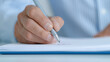 © TH_Stock - A close-up view of a hand holding a pen and writing on a sheet of paper. The background is softly blurred, emphasizing the act of writing and the details of the hand and pen.