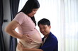 © kiattisak - Happy man with pregnant wife rubbing his belly while relaxing and spending time in modern apartment. Pregnant woman and husband spending time together using smartphone