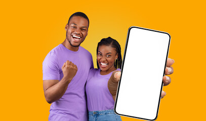 Fototapete - African American young couple, dressed in purple shirts, stand in front of a solid yellow background. The woman, holding a smartphone with a blank screen, points it towards the camera