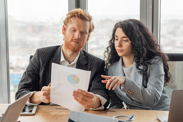 Aufkleber - Two colleagues, a man and a woman, are seated at a desk in a modern office. They are reviewing a document, likely a report with a pie chart, as part of a business meeting.